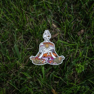 May include: A die-cut sticker featuring a person in a seated yoga pose. The design inside the silhouette includes a sun, mountains, flowers, and stars, with a color palette of orange, pink, and green. The sticker is placed on a bed of green grass.