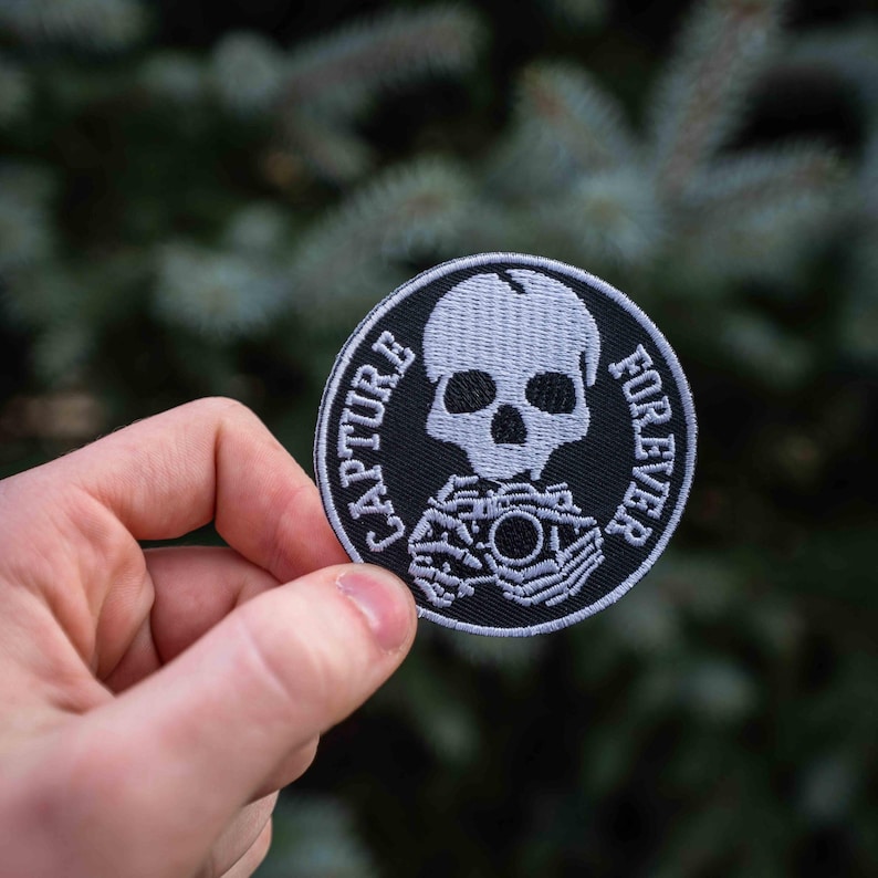 Pu&ograve; includere: Una toppa ricamata in bianco e nero con uno scheletro che tiene una macchina fotografica. Il testo "CAPTURE FOREVER" &egrave; scritto intorno allo scheletro.
