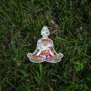 May include: A die-cut sticker in the shape of a person in a seated yoga pose. The sticker's interior features a colorful landscape with a sun, mountains, and flowers. The sticker is set against a backdrop of green grass.