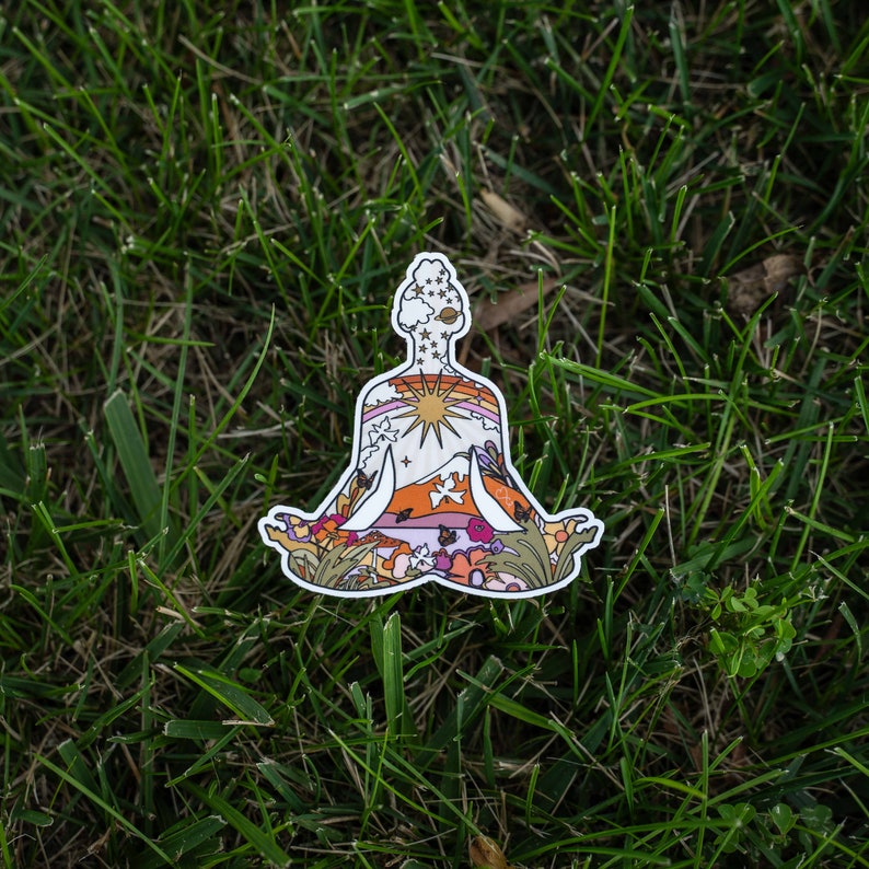 May include: A die-cut sticker in the shape of a person in a seated yoga pose. The sticker's interior features a colorful landscape with a sun, mountains, and flowers. The sticker is set against a backdrop of green grass.