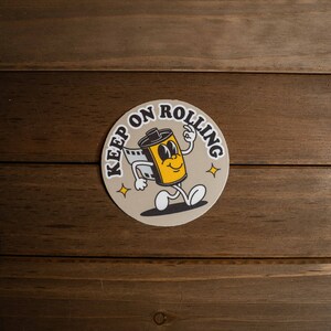 Puede incluir: Una pegatina redonda con un personaje de rollo de pel&iacute;cula de dibujos animados con el texto "Keep on Rolling" en letras marrones sobre un fondo beige.