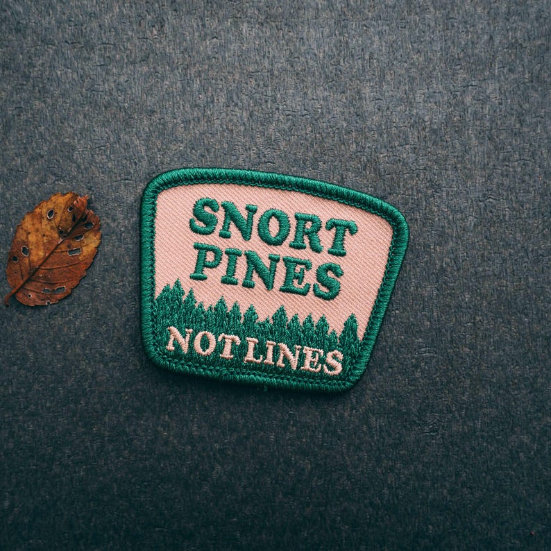 May include: A pink and green embroidered patch with the text "Snort Pines Not Lines".