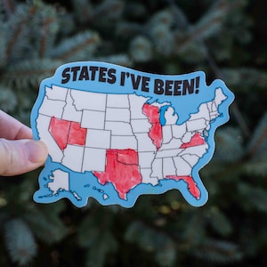 May include: A sticker in the shape of the United States with the text "STATES I'VE BEEN!" in blue. Several states are colored red. The sticker is held against a background of green foliage.