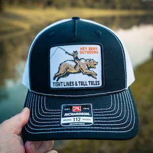 Könnte beinhalten: Eine schwarz-weiße Trucker-Cap mit einem rechteckigen Aufnäher, der eine Person beim Angeln auf einem Bären zeigt. Der Aufnäher enthält den Text HEY BEAR OUTDOORS und TIGHT LINES & TALL TALES. Ein schwarzer Aufkleber auf dem Schirm zeigt RICHARDSON 112.