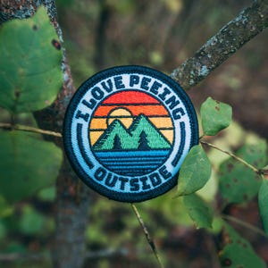 May include: A round, black and blue embroidered patch with the text "I Love Peeking Outside" and a retro sunset scene with mountains and trees.