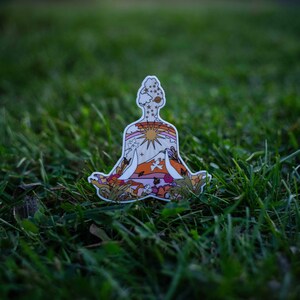 May include: A die-cut sticker in the shape of a person in a seated yoga pose. The sticker features a colorful design with a sun, rainbow, mountains, and flowers. The color palette includes orange, pink, yellow, and green.