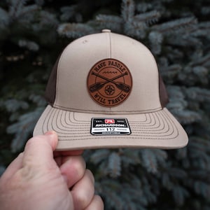 Könnte beinhalten: Eine braune und beigefarbene Trucker-Kappe mit einem braunen Leder-Patch, auf dem "Have Paddle Will Travel" mit einem Kompass und gekreuzten Paddeln steht. Die Kappe ist von Richardson 112.
