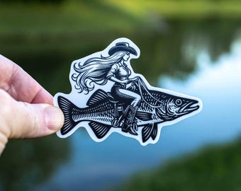 May include: Black and white sticker featuring a cowgirl riding a fish. The cowgirl wears a cowboy hat, shirt, and boots. The fish has detailed scales and fins. The sticker is held against a blurred green and blue background.