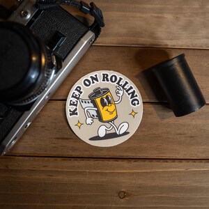 Puede incluir: Un rollo de pel&iacute;cula de dibujos animados amarillo y negro con una cara sonriente y el texto "Keep on Rolling" en un c&iacute;rculo beige.