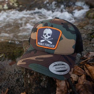 May include: A camouflage trucker hat with a black mesh back. The hat has a patch with a skull and crossbones and the text "Paddle Forever". The hat also has a silver sticker that says "The Authentic Snapback Pupdoong Since 1974".