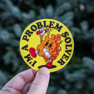 以下が含まれることがあります： 黒い文字で「I'M A PROBLEM SOLVER」と書かれた丸い黄色のステッカー。赤い靴を履き、笑顔の顔をした、火の玉のキャラクターが点火された爆竹を持っている様子が描かれています。