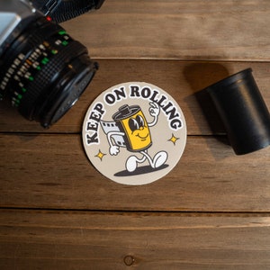 Puede incluir: Un posavasos redondo de color beige con el texto "Keep on Rolling" en letras negras. Un rollo de pel&iacute;cula de dibujos animados con una cara sonriente camina por el posavasos.
