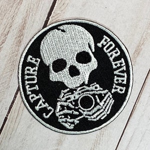 Pu&ograve; includere: Toppa ricamata in bianco e nero con uno scheletro che tiene una macchina fotografica. Il testo "Capture Forever" &egrave; scritto intorno allo scheletro.