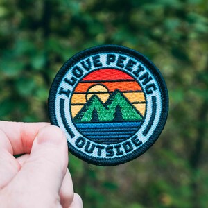 May include: A round, embroidered patch with a sunset and mountains design. The patch is white with black and blue accents and says "I Love Peeking Outside".