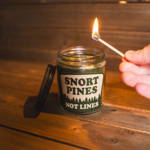 May include: A green glass candle jar with a white label that reads "Snort Pines Not Lines". The candle is being lit with a match.