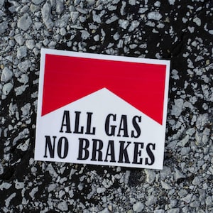 Könnte beinhalten: Ein rechteckiger Aufkleber mit einem rot-weißen Chevron-Muster und dem schwarzen Text ALL GAS NO BRAKES in einer fetten Serifenschrift. Der Aufkleber hat einen sauberen weißen Rand und liegt auf einer strukturierten grauen und schwarzen Felsfläche.