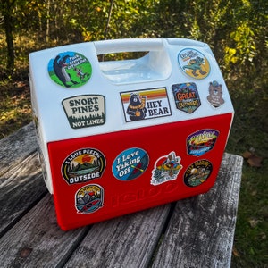 May include: A red and white Igloo cooler covered in outdoor-themed stickers. The stickers feature phrases like "Explore More," "Snort Pines Not Lines," and "I Love Peeing Outside." The cooler sits on a weathered wooden surface.