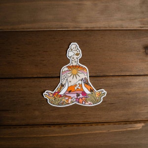 May include: A die-cut sticker featuring a person in a seated yoga pose. The silhouette is filled with a colorful design of a sun, rainbow, mountains, flowers, and butterflies. The sticker has a white border and is set against a wooden background.