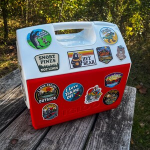 May include: A red and white Igloo cooler adorned with various outdoor adventure stickers. The stickers display slogans such as "Explore More," "Snort Pines Not Lines," and "I Love Peeing Outside." The cooler is placed on a wooden surface.
