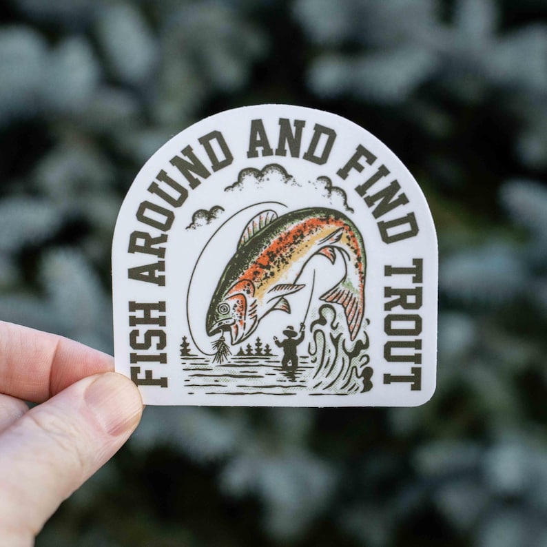 K&ouml;nnte beinhalten: Wei&szlig;er Aufkleber mit dem Text "FISH AROUND AND FIND TROUT" in einem Halbkreis. Der Aufkleber zeigt eine springende Forelle mit orangefarbenen, roten und gr&uuml;nen Details, einen Fischer und eine Waldszene.