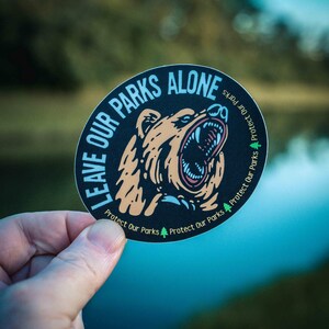 May include: A circular sticker with the text "LEAVE OUR PARKS ALONE" and "Protect Our Parks". The sticker features a roaring bear illustration in shades of brown, black, and blue. The sticker is held by a hand against a blurred natural background.