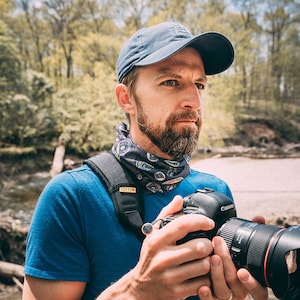 May include: A man wearing a blue t-shirt, a blue baseball cap, and a patterned neck gaiter holds a black Canon camera with a lens. He is standing in a wooded area near a stream.