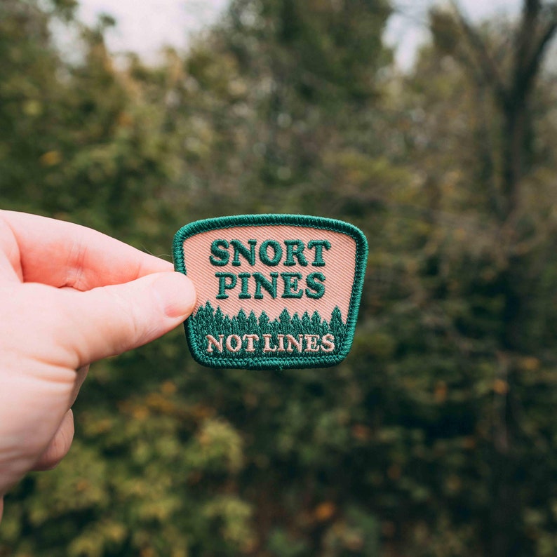 May include: A pink and green embroidered patch with the text "Snort Pines Not Lines".