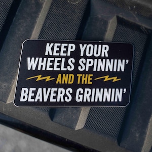 Könnte beinhalten: Ein rechteckiger schwarzer Aufkleber mit weißem Text: "KEEP YOUR WHEELS SPININ' AND THE BEAVERS GRINNIN'". Zwei gelbe Blitzsymbole sind ebenfalls abgebildet.