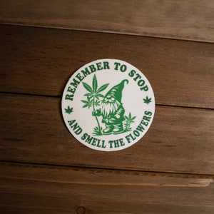 May include: White round sticker with green text that reads "Remember to Stop and Smell the Flowers". A green cartoon gnome holding a green marijuana plant is in the center of the sticker.