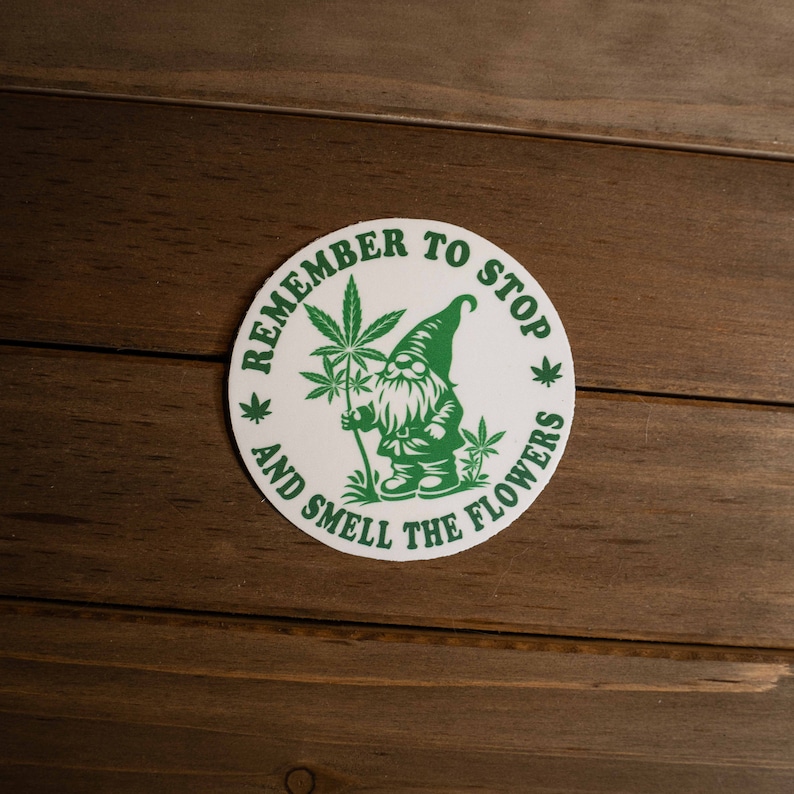 May include: White round sticker with green text that reads "Remember to Stop and Smell the Flowers". A green cartoon gnome holding a green marijuana plant is in the center of the sticker.