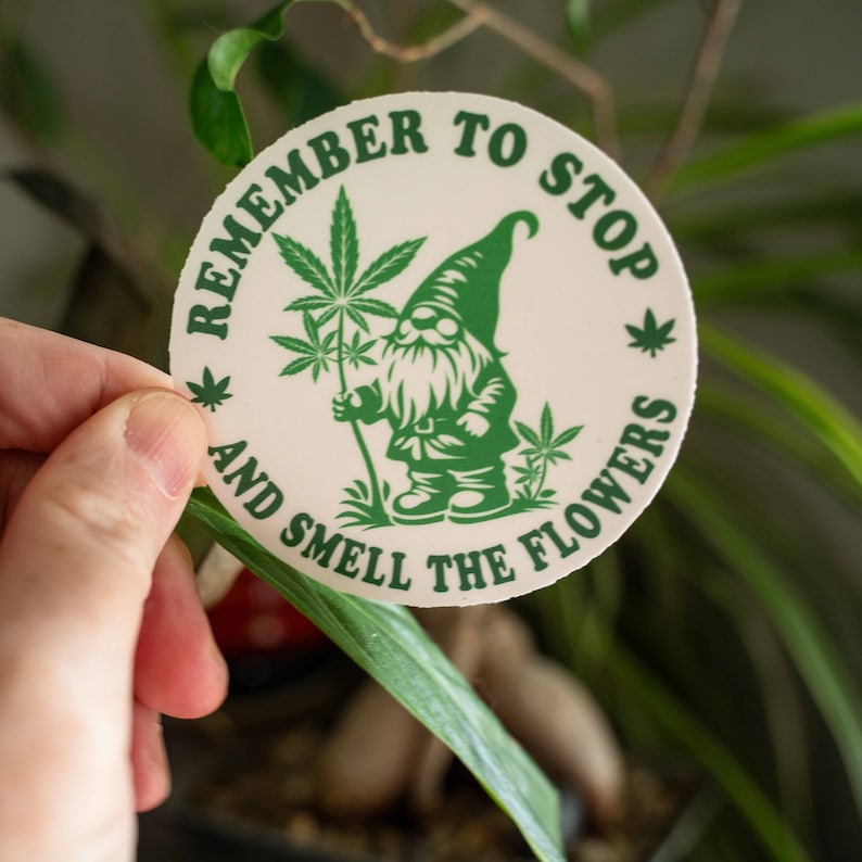 May include: A white round sticker with green text and a green cartoon gnome holding a marijuana plant. The text reads "Remember to stop and smell the flowers".