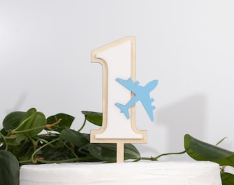 May include: A white wooden number one cake topper with a blue airplane on top. The cake topper is on a white cake.