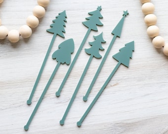 May include: Six teal green acrylic drink stirrers shaped like Christmas trees. The stirrers are on a white wooden surface.
