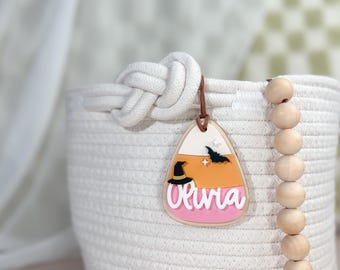 May include: A white rope basket with a candy corn shaped tag that says "Olivia". The tag has a witch's hat, bats, and stars on it. The basket is decorated with wooden beads.