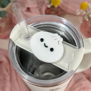 May include: White stainless steel travel mug with a white lid and a white handle. The lid has a white silicone ring and a white plastic straw. The mug has a white plastic charm with a smiling face on it.