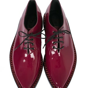 K&ouml;nnte beinhalten: Ein Paar bordeauxfarbene Lacklederschuhe mit schwarzen Schn&uuml;rsenkeln. Die Schuhe haben eine spitze Zehenpartie und eine Kettenverzierung um die Sohle. Gr&ouml;&szlig;e 42 (EU).