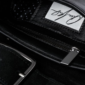 May include: Black leather belt with a silver buckle and a zipped pocket. The pocket has a black leather lining and a white label with the text "Carry Away" written in black.