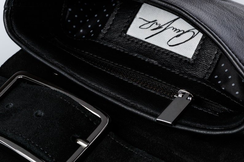May include: Black leather belt with a silver buckle and a zipped pocket. The pocket has a black leather lining and a white label with the text "Carry Away" written in black.