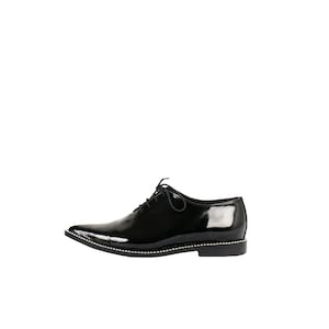 K&ouml;nnte beinhalten: Ein einzelner schwarzer Lacklederschuh mit spitzer Zehenpartie und Schn&uuml;rverschluss. Der Schuh hat eine dekorative Reihe kleiner silberner Nieten entlang des Sohlenrandes. Geeignet f&uuml;r formelle Anl&auml;sse.
