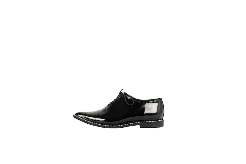 K&ouml;nnte beinhalten: Ein einzelner schwarzer Lacklederschuh mit spitzer Zehenpartie und Schn&uuml;rverschluss. Der Schuh hat eine dekorative Reihe kleiner silberner Nieten entlang des Sohlenrandes. Geeignet f&uuml;r formelle Anl&auml;sse.
