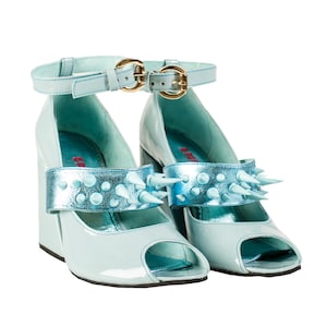 May include: A pair of light blue peep-toe heels with a wedge heel. The shoes feature a metallic blue strap with light blue spikes and a matching ankle strap with gold buckles. The shoes have a glossy finish.