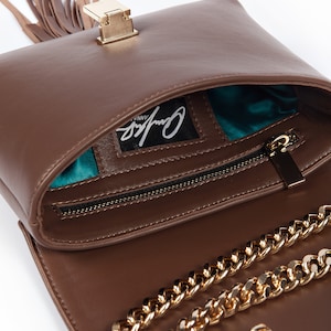 May include: A brown leather handbag with a gold chain strap and a gold clasp. The bag has a tassel and a teal lining. The bag has the brand name "Cynthia Vinny" printed on the inside.