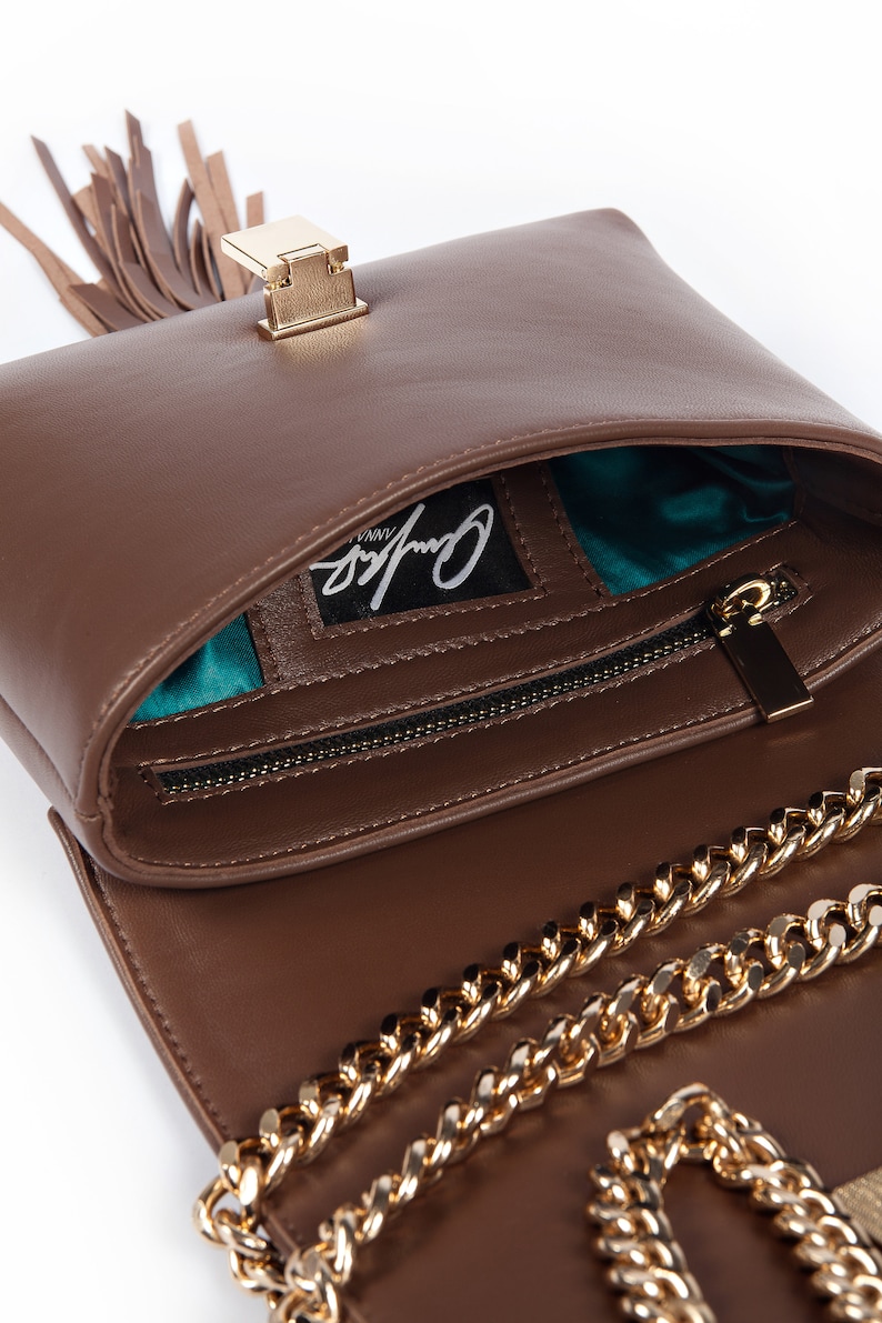 May include: A brown leather handbag with a gold chain strap and a gold clasp. The bag has a tassel and a teal lining. The bag has the brand name "Cynthia Vinny" printed on the inside.