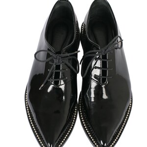 K&ouml;nnte beinhalten: Ein Paar schwarze Lacklederschuhe mit spitzer Zehenpartie. Die Schuhe haben einen Schn&uuml;rverschluss und sind mit kleinen silberfarbenen Nieten besetzt.