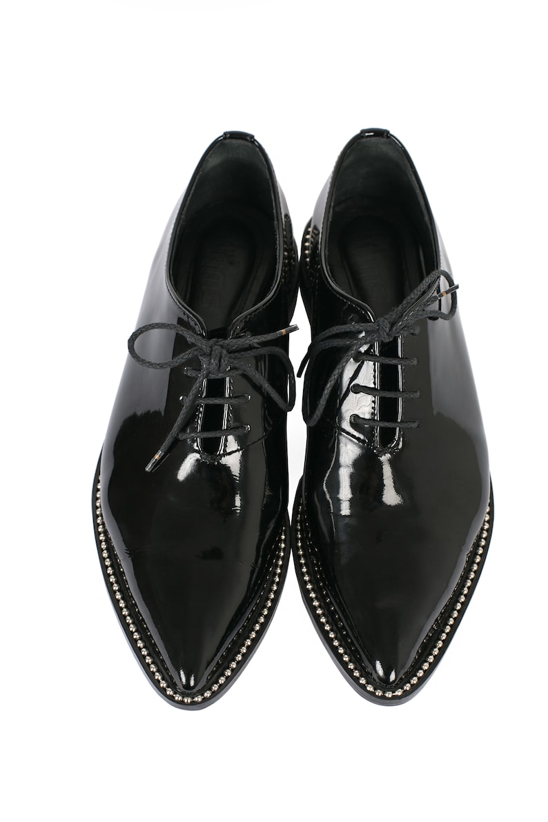 K&ouml;nnte beinhalten: Ein Paar schwarze Lacklederschuhe mit spitzer Zehenpartie. Die Schuhe haben einen Schn&uuml;rverschluss und sind mit kleinen silberfarbenen Nieten besetzt.