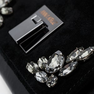 May include: A black suede clutch bag with a silver clasp and the text "ANNA KRUZ" on the front. The bag is adorned with several clear crystal embellishments.