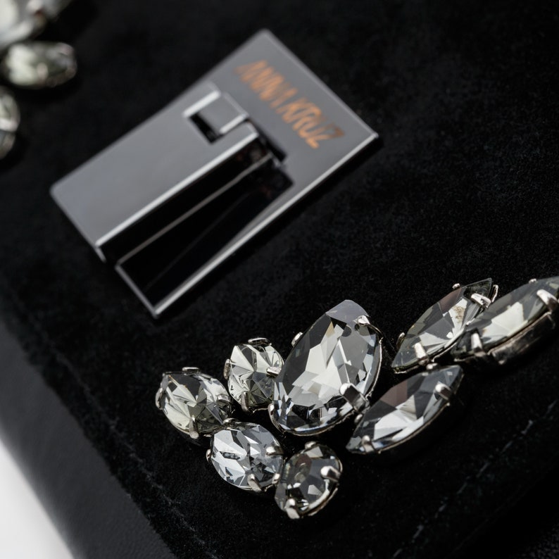 May include: A black suede clutch bag with a silver clasp and the text "ANNA KRUZ" on the front. The bag is adorned with several clear crystal embellishments.