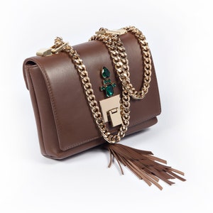 May include: A brown leather shoulder bag with a gold chain strap and a green gem embellishment. The bag has a tassel hanging from the bottom.