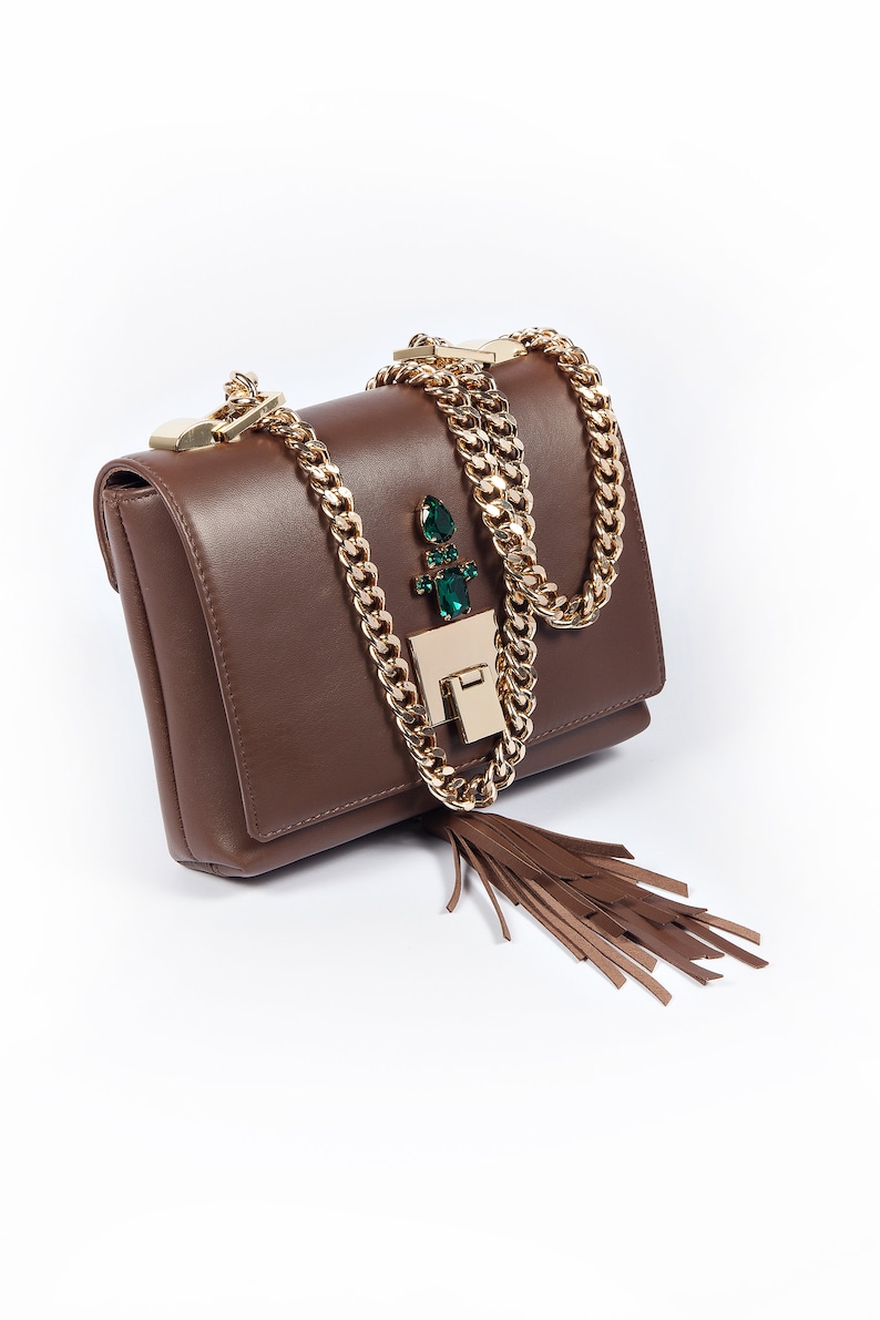 May include: A brown leather shoulder bag with a gold chain strap and a green gem embellishment. The bag has a tassel hanging from the bottom.