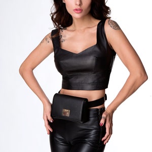 May include: A woman wearing a black leather crop top and matching pants with a black fanny pack. The fanny pack has a gold buckle.